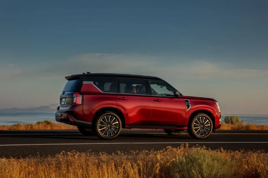 Angled right profile view of a red 2025 Nissan Armada parked on a brush lined road at dusk with a body of water behind.