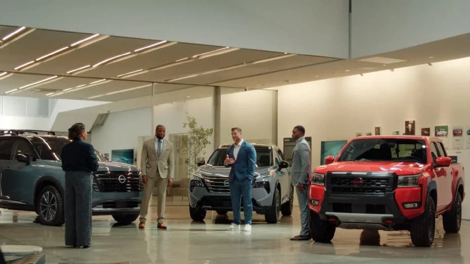 A group of people in business clothes, standing around three Nissan vehicles in a showroom.