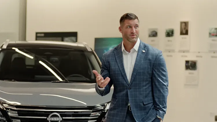 Tim Tebow in a plaid suit, standing in front of a Nissan Rogue, holding his hand up and looking to the right.